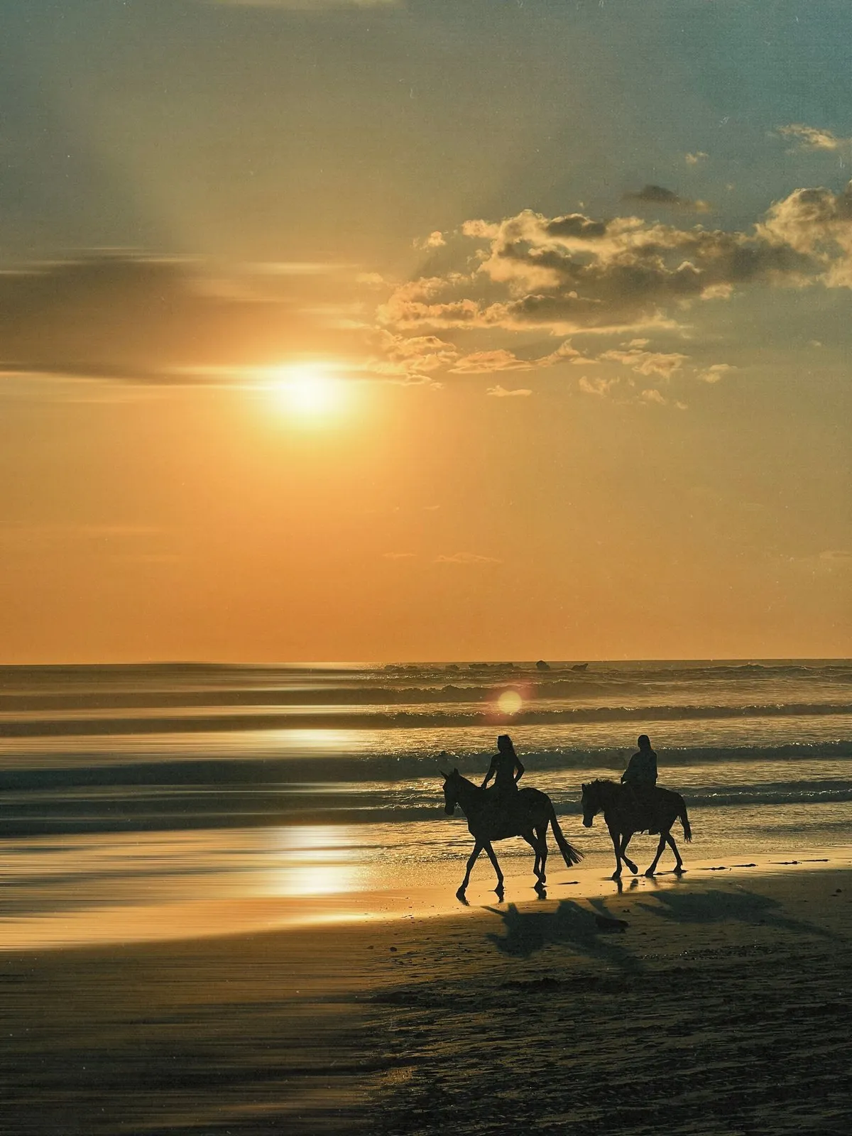 Two riders on horseback along the beach at sunset